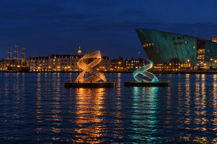 Two helix light sculptures on water at night with cityscape in background.