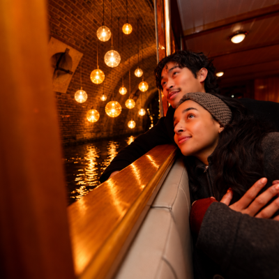 Couple gazes out boat window at night, warm lights reflect on water.