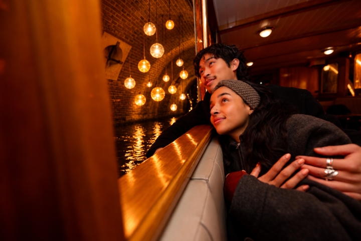 Couple gazes out boat window at night, warm lights reflect on water.